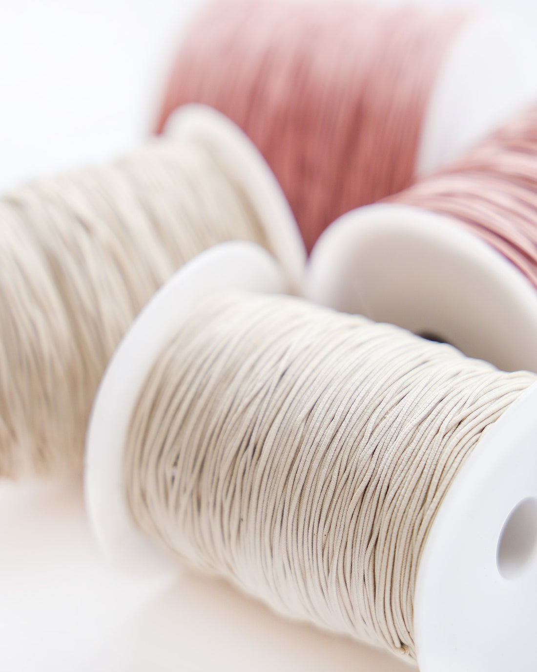 Nahaufnahme von drei Garnspulen in Creme-, Rosa- und Beigetönen, mit Weichzeichner-Hintergrund.