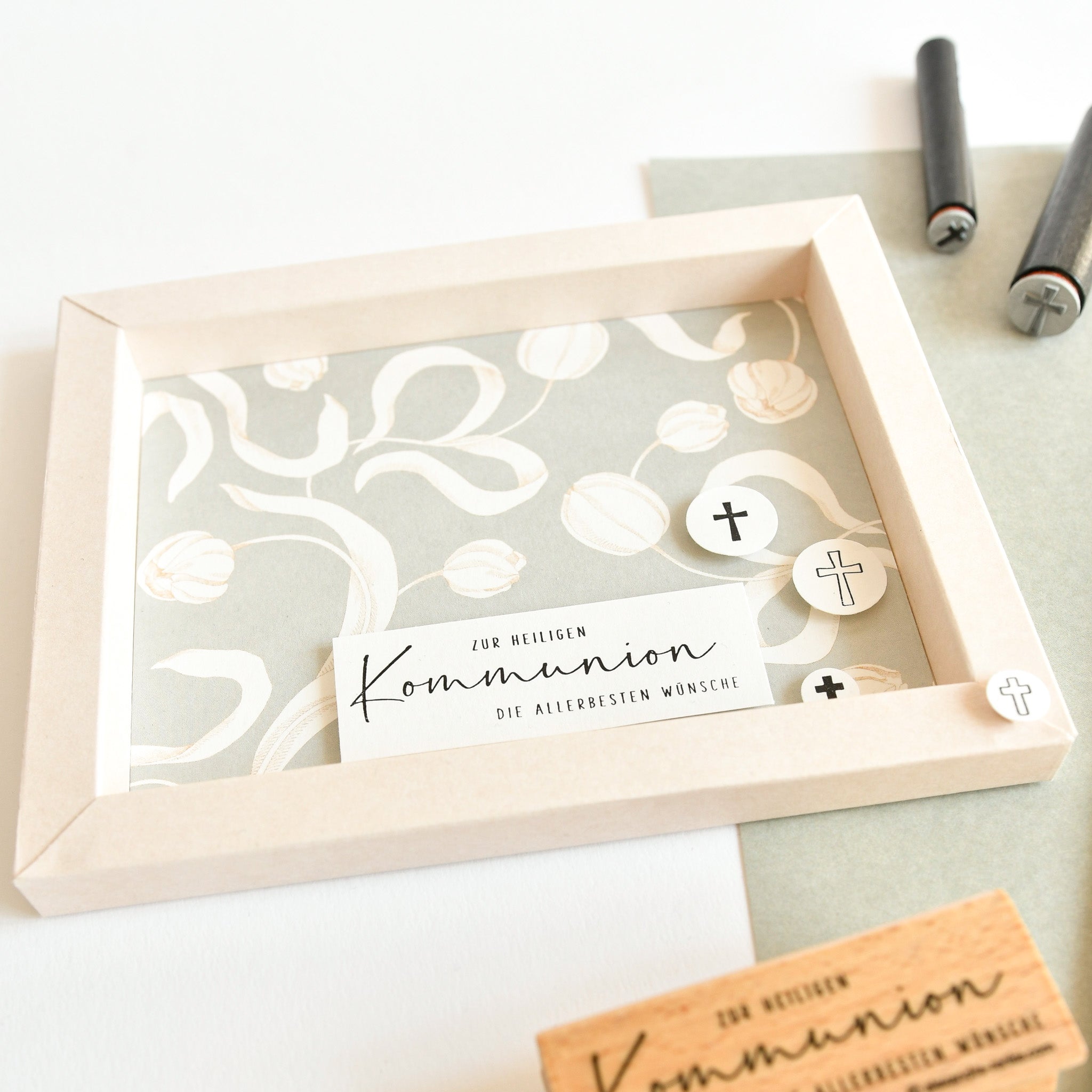 Gerahmte Kommunionskarte mit Blumenmuster und Kreuzsymbolen, mit deutschem Text und einem Mini Kreuz Holzstempel von Alexandra Renke, auf einem hellen Schreibtisch mit Stiften in der Nähe.