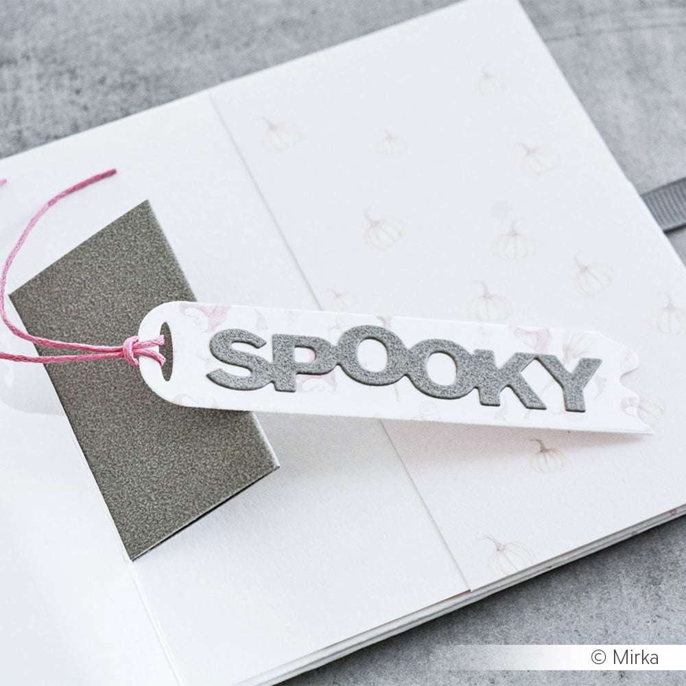 Ein weißes Lesezeichen von Alexandra Renke mit dem grauen Schriftzug SPOOKY" und der Motivstanze Etikett Spooky liegt auf einem aufgeschlagenen Notizbuch, an dem eine rosa Schnur befestigt ist.