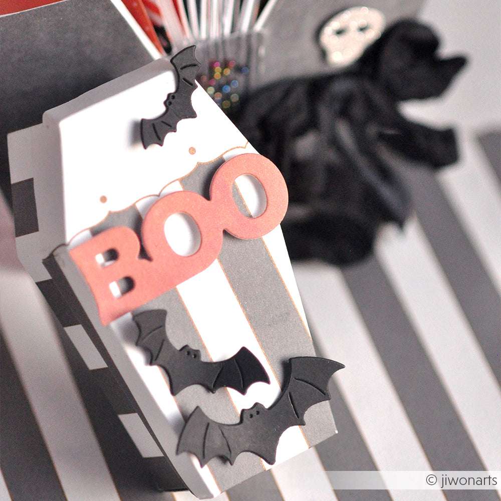 Schwarz-weiße Schachtel in Sargform mit "BOO" und Fledermäusen, hergestellt mit den DieCut Systemen von Alexandra Renke und der Motivstanze Fledermausschwarm, umgeben von Halloween-Dekorationen im Hintergrund.