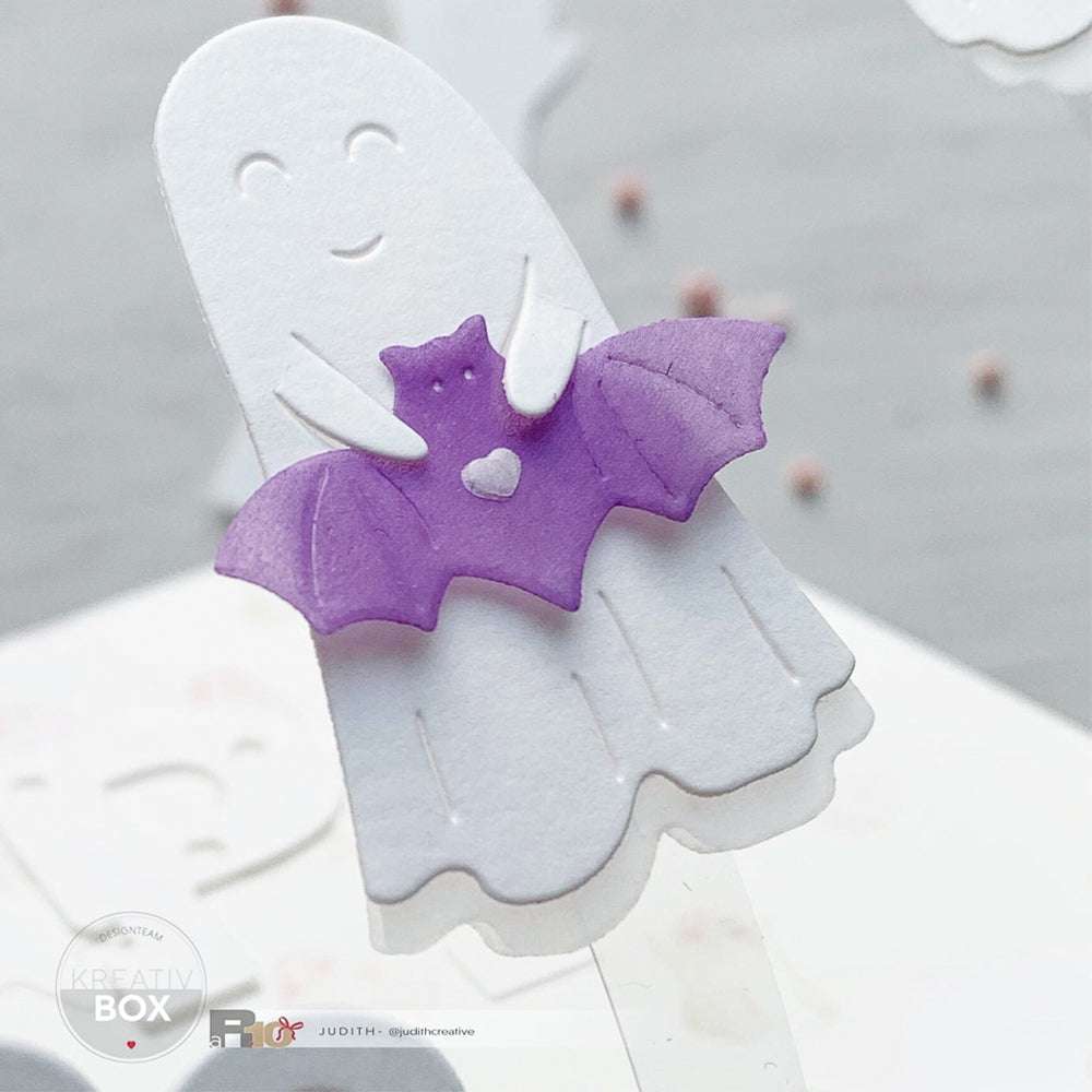 Ein weißes Papiergespenst hält eine kleine lila Fledermaus mit einem Herz auf der Brust; beide sind aus Alexandra Renke Designpapieren mit kreativen Stanzsystemen hergestellt.