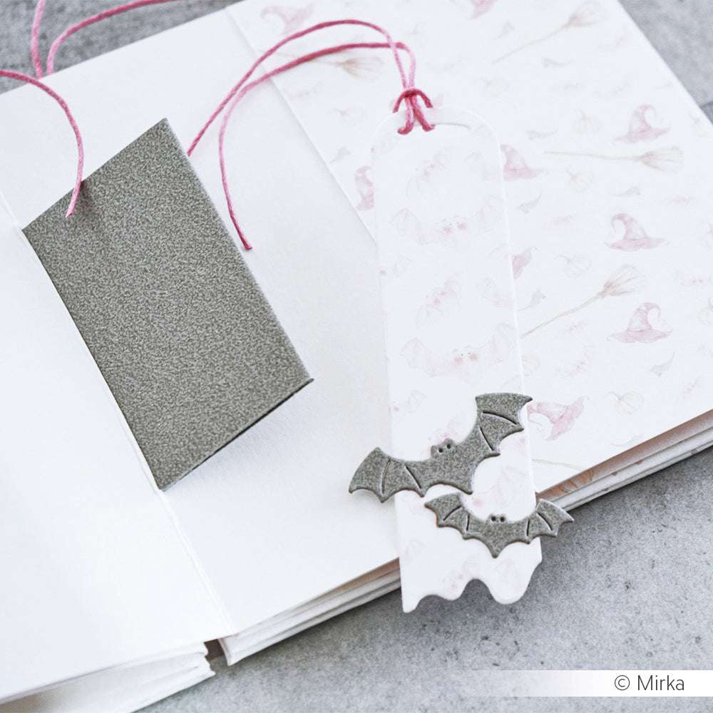 Ein weißes Lesezeichen mit Fledermausmotiv, hergestellt mit Alexandra Renke Motivstanze Fledermausschwarm und rosa Kordel, ruht auf einem aufgeschlagenen Notizbuch mit pastellfarbenen Fledermausmotiven.