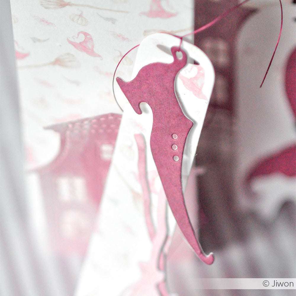 Ein rosafarbenes Papierornament in Form eines Seepferdchens, das mit einer Alexandra Renke DieCut Systeme Stanzen Motivstanze Hexenzubehör hergestellt wurde, hängt vor einem sanft verwischten gemusterten Hintergrund.