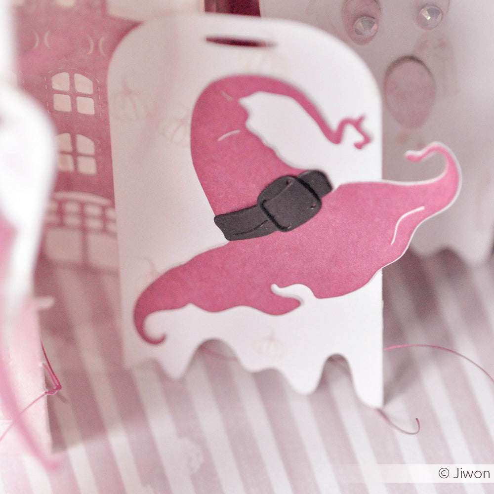 Ein rosa Hexenhut mit schwarzem Gürtel ist auf einem weißen, geisterförmigen Anhänger mit gestreiftem Hintergrund abgebildet - ideal zur Kombination mit Alexandra Renke Designpapieren oder zum Basteln mit der Motivstanze Hexenzubehör.