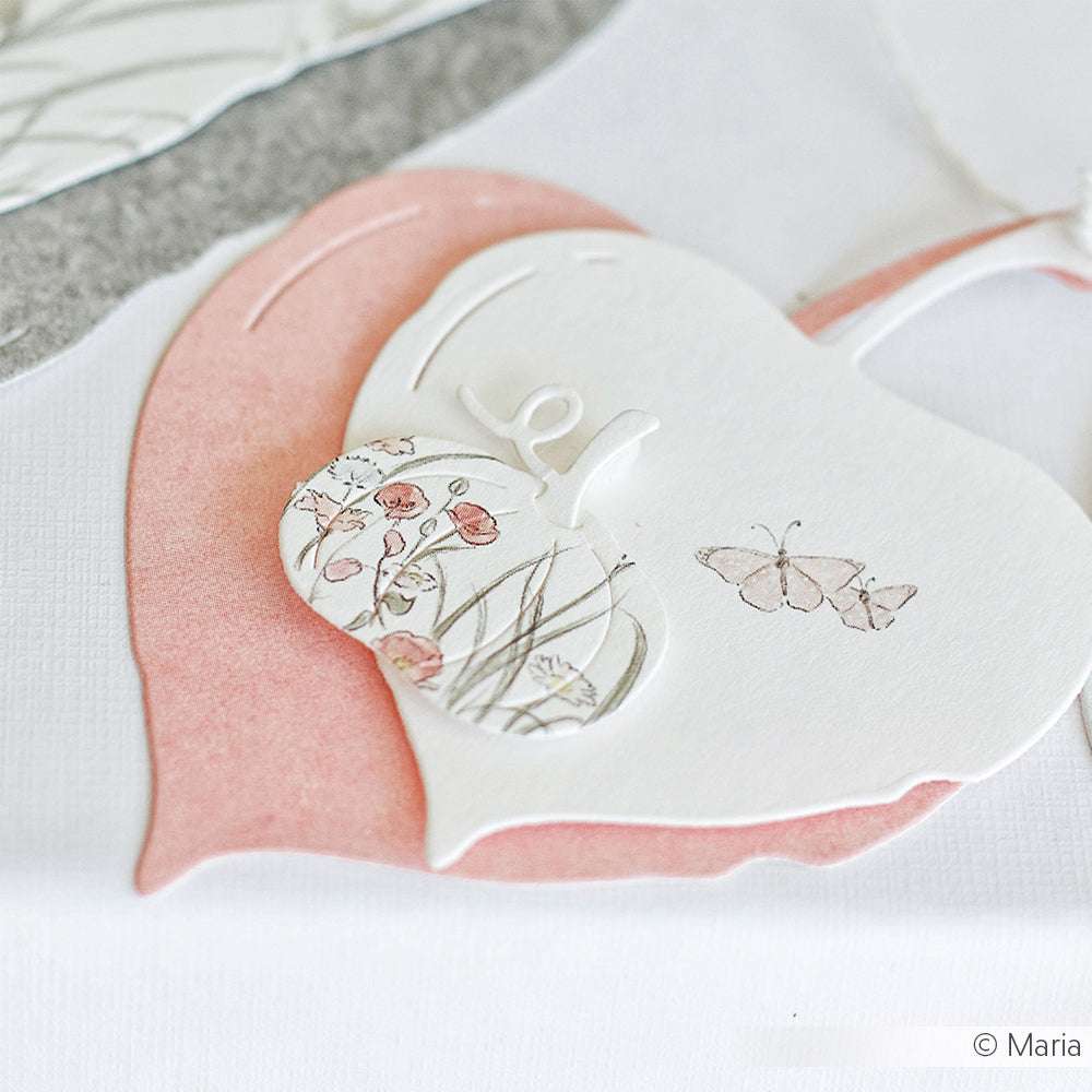 Papierherzen mit Blumen- und Schmetterlingsmotiven, die mit den Stanzformen von Alexandra Renke hergestellt wurden, sind wunderschön auf einer weißen Oberfläche geschichtet.