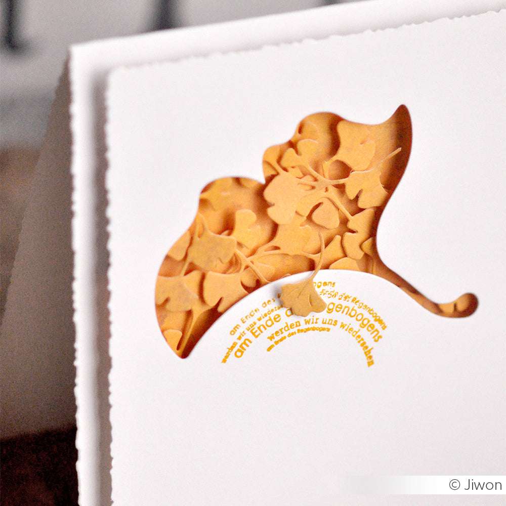 Nahaufnahme einer mit Alexandra Renke Designpapieren hergestellten Papierkarte mit einem Motivstanze Ginkoblätter-Ausschnitt, der gelben Text und Blattformen im Inneren zeigt.