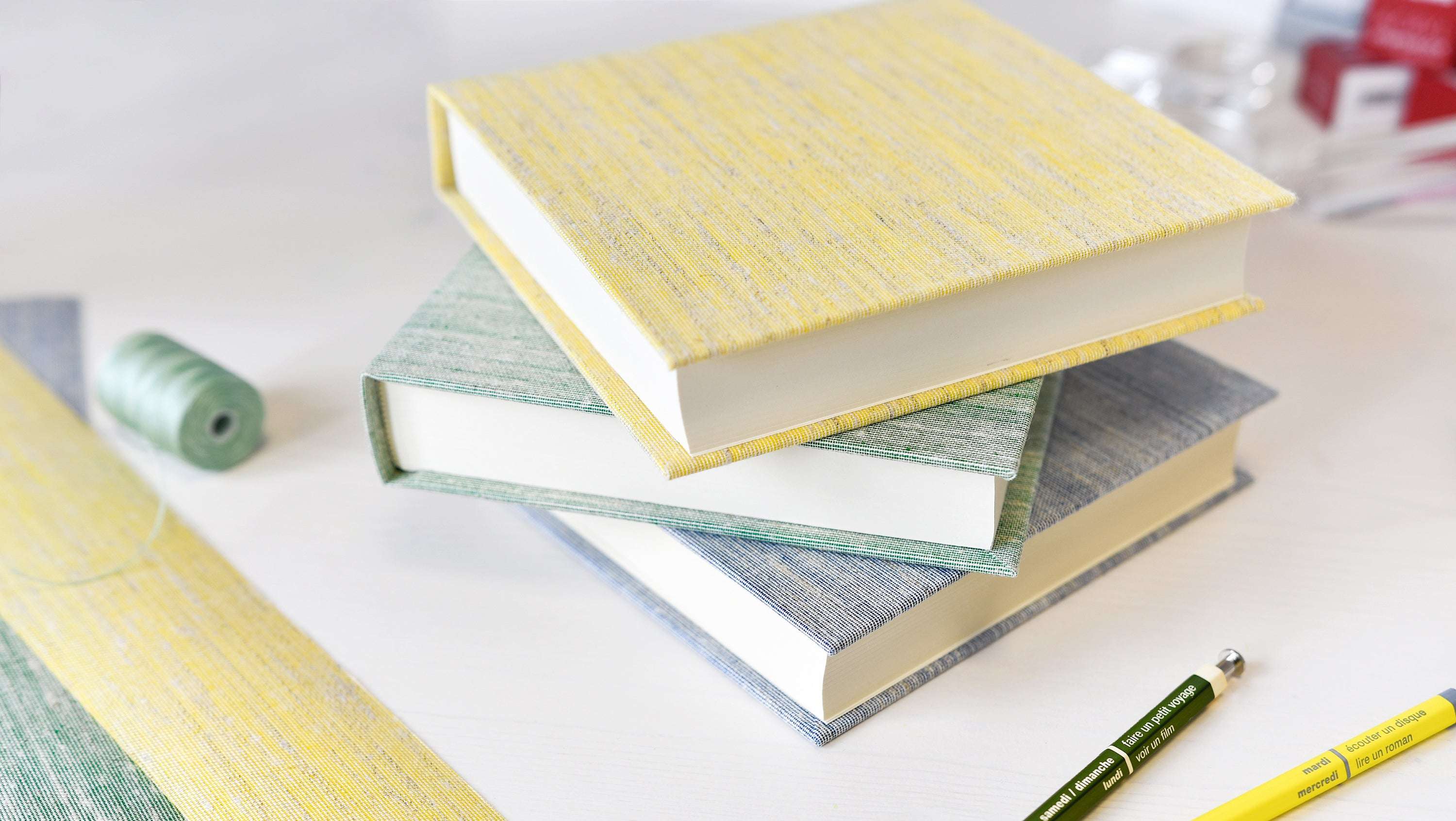 Drei gebundene Bücher mit pastellfarbenem Einband liegen auf einem weißen Schreibtisch, umgeben von Schreibwaren.
