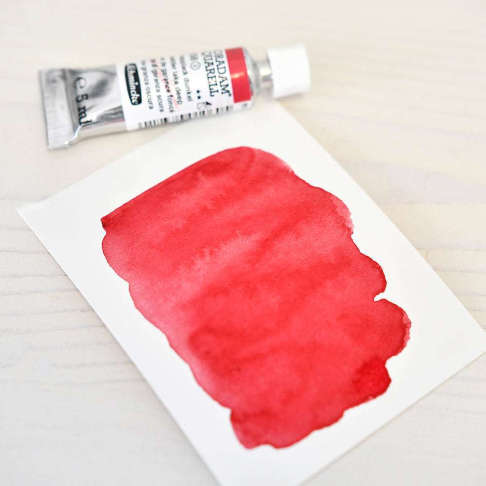 Eine Tube Schmincke-Aquarellfarbe liegt neben einem Blatt, das ein rotes Muster auf einer hellen Holzfläche zeigt.