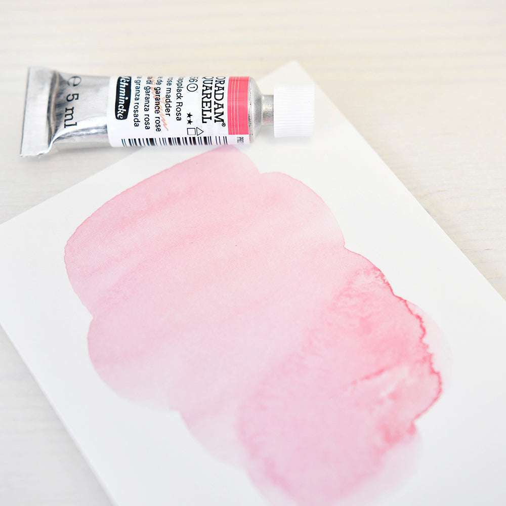 Eine Tube Schmincke-Aquarellfarbe in Krapplack Rosa steht neben einem Blatt, auf dem Künstler-Aquarellfarben in Rosa auf einer hellen Fläche verwischt sind.