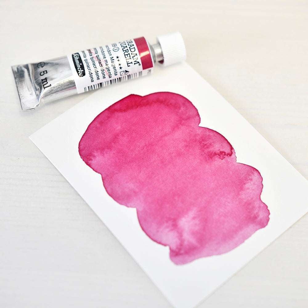 Eine Tube Schmincke Aquarellfarbe in Chinacridon Magenta liegt neben einer Karte, die ein rosa Muster auf einer weißen Fläche zeigt.