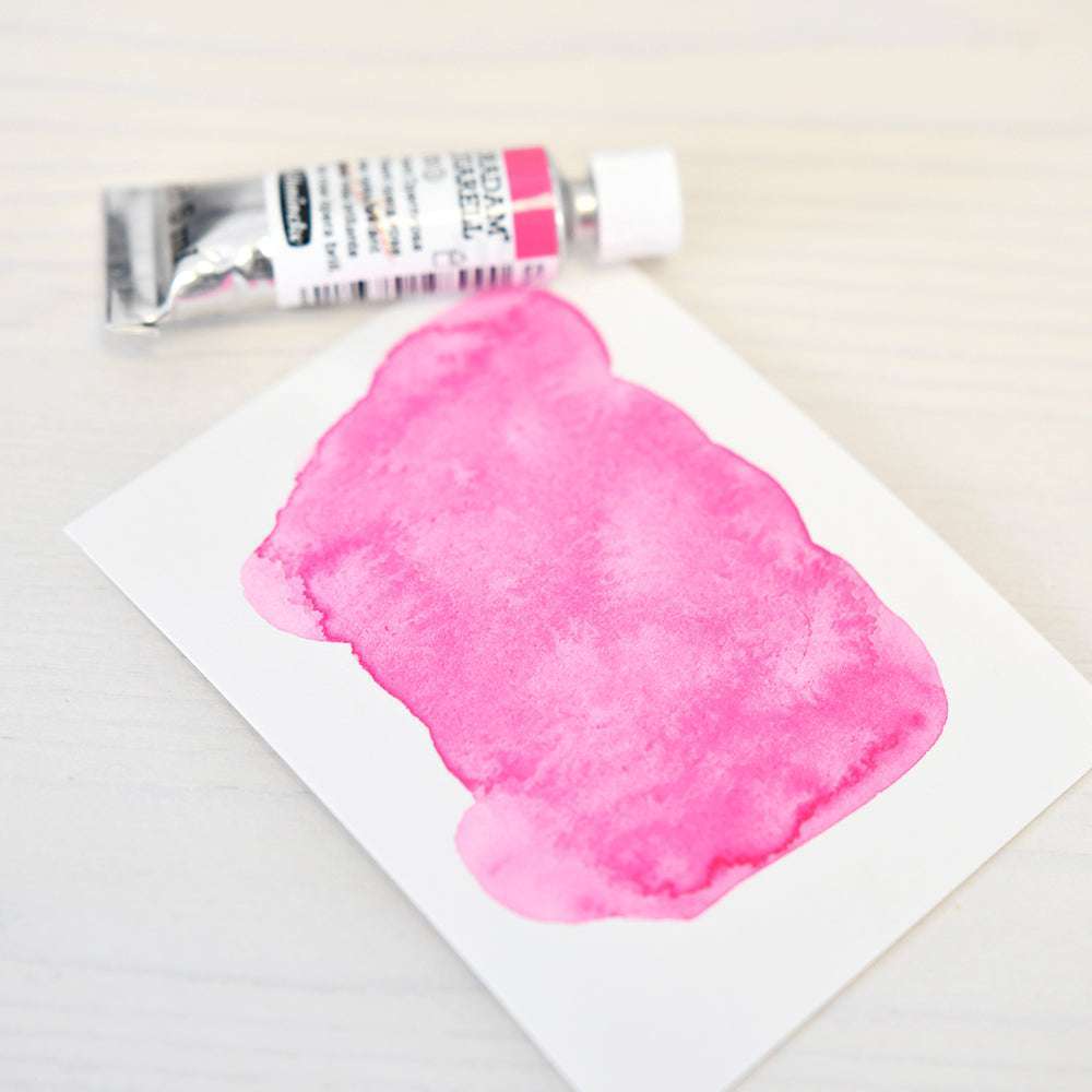 Eine Tube Schmincke-Aquarellfarbe in Brilliant Opera Rose liegt neben einer Karte mit einem kräftigen rosa Farbtupfer, beide auf einer weißen Fläche.