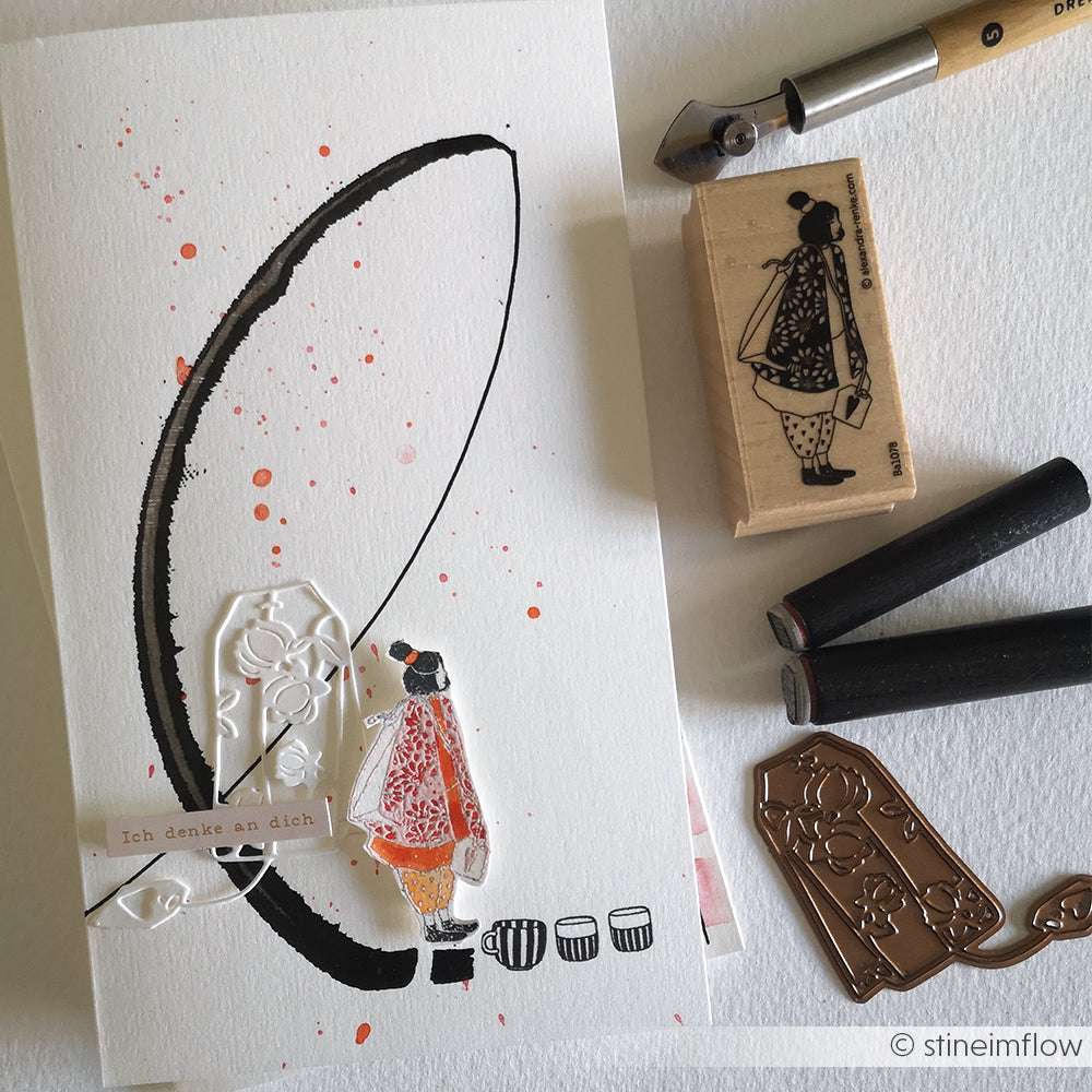 Handgefertigte Karte mit gestempeltem Mädchenmotiv, Farbspritzern und Alexandra Renke-Bastelwerkzeugen wie Holzstempel und Akzent-Stempel auf einer weißen Fläche.