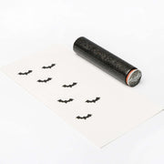 Ein Holzstempel in Fledermausform von Alexandra Renke erzeugt einen charmanten Effekt, indem er mehrere schwarze Fledermausabdrücke auf weißes Papier stempelt und so einen auffälligen Akzent setzt.