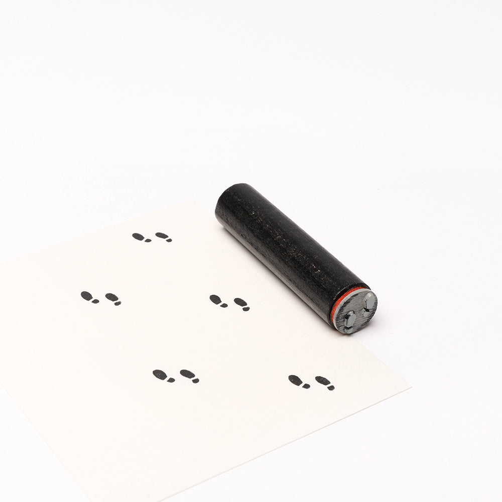 Ein schwarzer Alexandra Renke Stempel Roller druckt Mini Fußspuren als kleine schwarze Abdrücke auf ein weißes Blatt Papier.