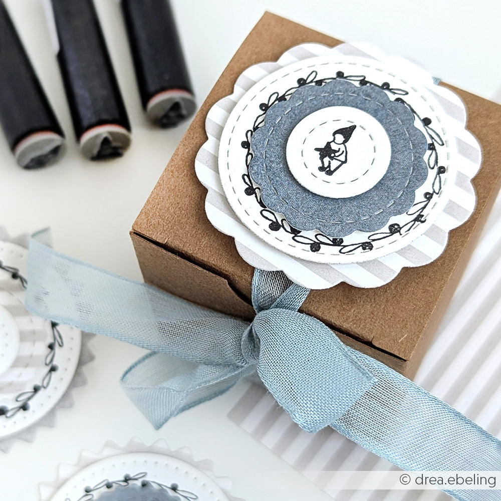 Kleine braune Geschenkschachtel mit blauem Band und einem runden, geschichteten Anhänger mit der Illustration Mini Wichtel mit Stern von Alexandra Renke, erstellt mit einem Akzent-Stempel.