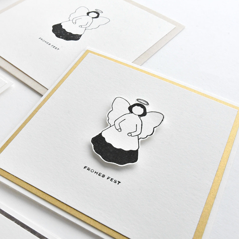 Quadratische Grußkarte von Alexandra Renke mit einer Schwarz-Weiß-Illustration von Fröhlicher Engel, die mit einem hochwertigen Stempel Holz hergestellt wurde, und dem darunter gedruckten "Frohes Fest".