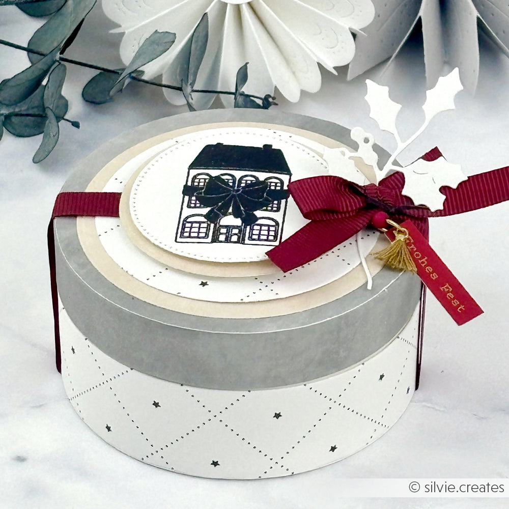 Runder weißer Alexandra Renke Geschenkkarton mit einer Hausillustration, rotem Band und Stechpalme auf Marmor - perfekt für die Feiertage mit seinem festlichen Schleifenhaus-Design, ideal zu kombinieren mit Stempelgummi oder Holzstempel-Kreationen.
