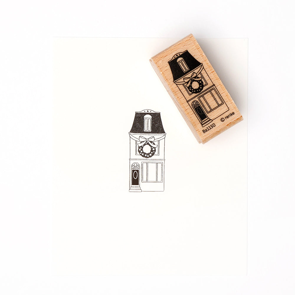 Ein hölzerner Alexandra Renke Holzstempel, der ein Haus mit einem Kranz an der Tür zeigt, steht auf einem weißen Hintergrund, der das detaillierte Stempelgummi hervorhebt.