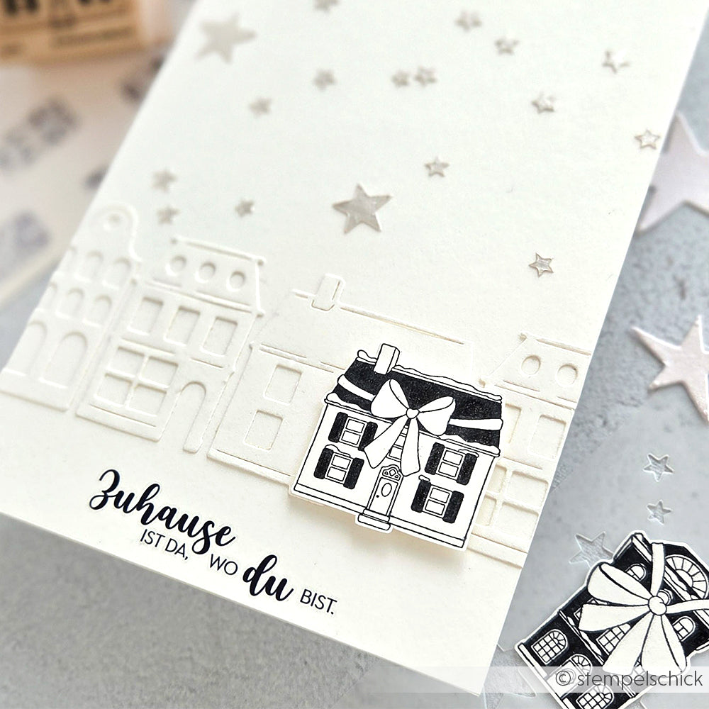 Eine handgefertigte Karte nach den Entwürfen von Alexandra Renke zeigt geprägte Häuser, einen schwarz-weißen Hausausschnitt, silberne Sterne, einen deutschen Text am unteren Rand und kreative Details, die von Papierprojekten und Stempelgummi inspiriert sind.