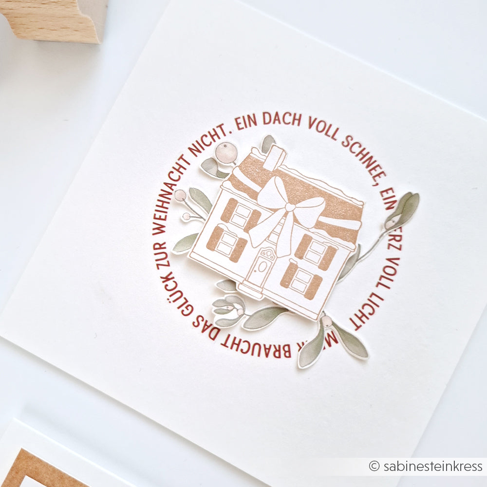 Eine Weihnachtskarte von Alexandra Renke mit einer Haus-Illustration, Holzstempel-gestempelt und umrahmt mit deutschem Text in einem roten Kreis auf weißem Papierprojekte.