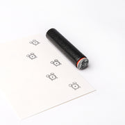 Ein schwarzer Alexandra Renke Holzstempel und ein Blatt Papier auf weißem Grund, das mit kleinen, einfachen Bärengesichtern bestempelt ist.