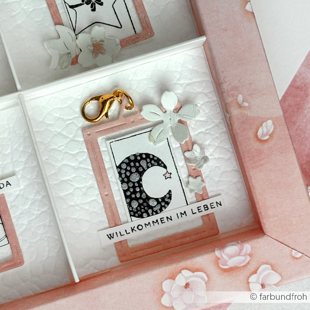 Eine rosa-weiße Schattenboxkarte von Alexandra Renke mit dem Buchstaben C, einem Stern, Blumen, dem Text "WILLKOMMEN IM LEBEN" und einem eleganten Holzstempel-Akzent.