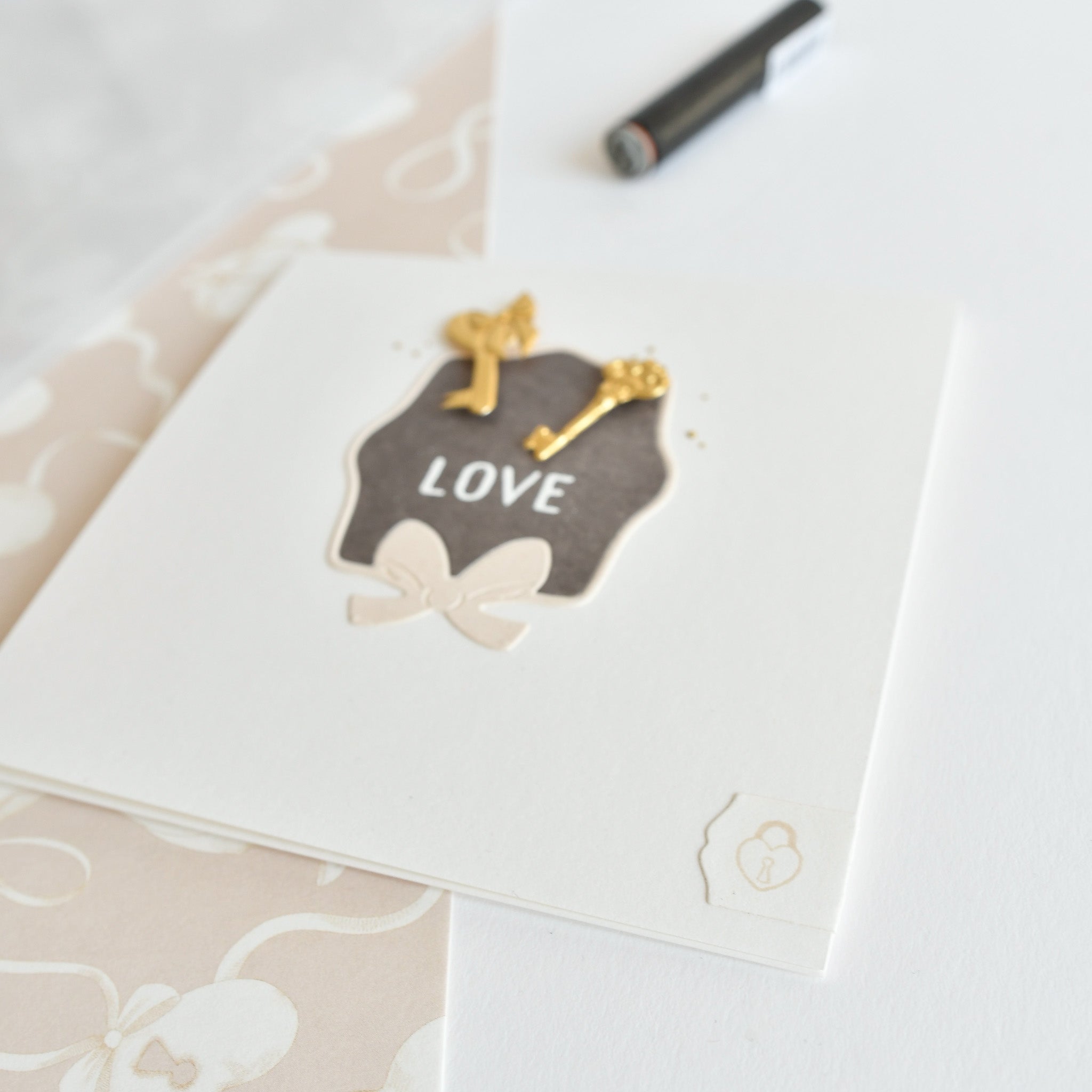 Quadratische Karte von Alexandra Renke mit "LOVE" und zwei goldenen Schlüsseln auf einem schwarzen Etikett, verziert mit einer Schleife und Mini-Herzensschloss, auf einer weißen Fläche.