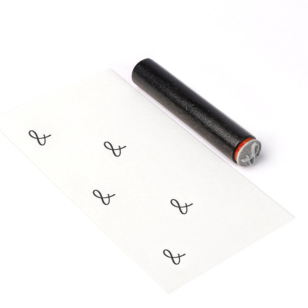 Eine schwarze Holzstempelwalze von Alexandra Renke stempelt wiederholte L-ähnliche Symbole in Reihen auf weißes Papier.