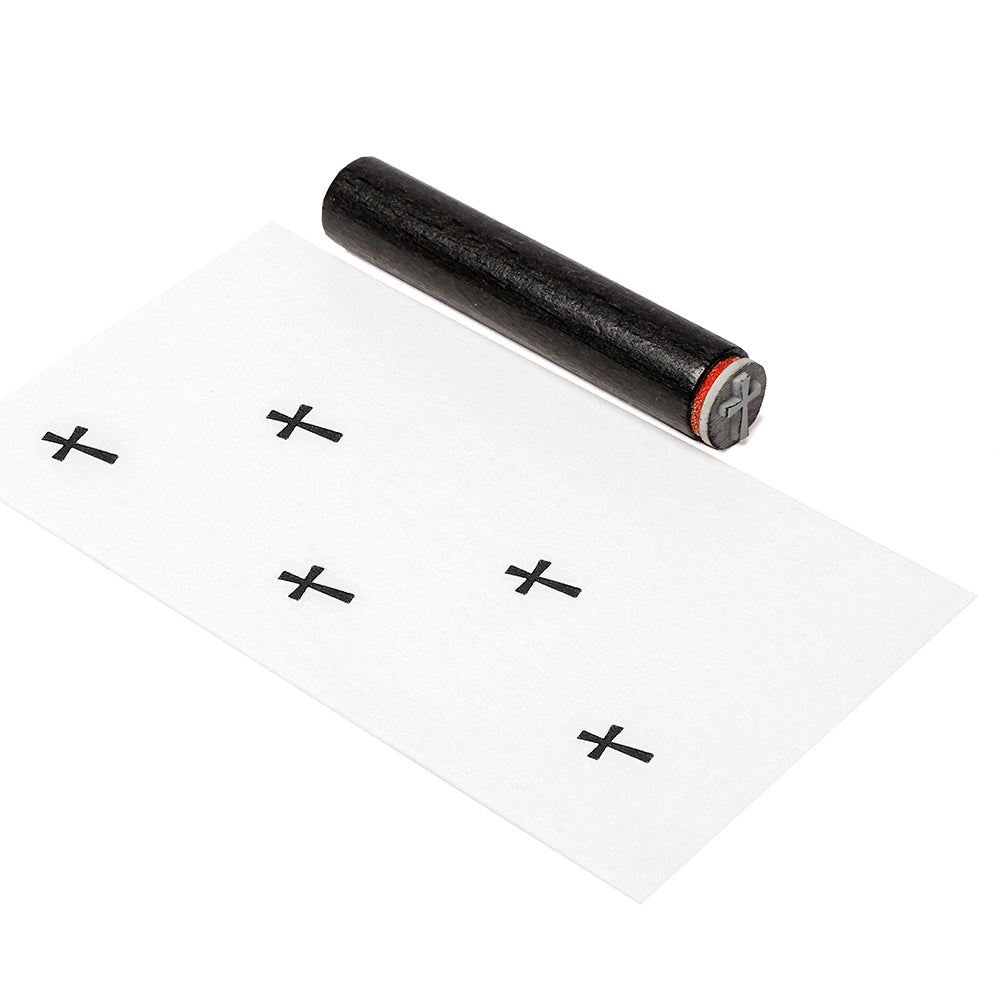 Ein schwarzer Holzstempel von Alexandra Renke mit einem Kreuzsymbol ruht neben gestempelten Kreuzen auf einem weißen Blatt und setzt klare Akzente.