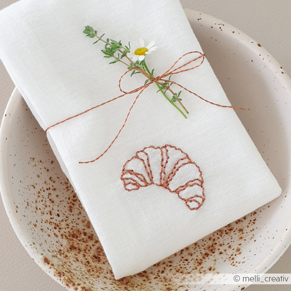 Eine weiße Alexandra Renke-Serviette mit einem gestickten Hörnchen, gebunden mit Bindfaden und Kräutern in einer gesprenkelten Keramikschale - ideal, um Ihr Sticken Zubehör oder inspirierende Stickvorlagen Stempel zu präsentieren.
