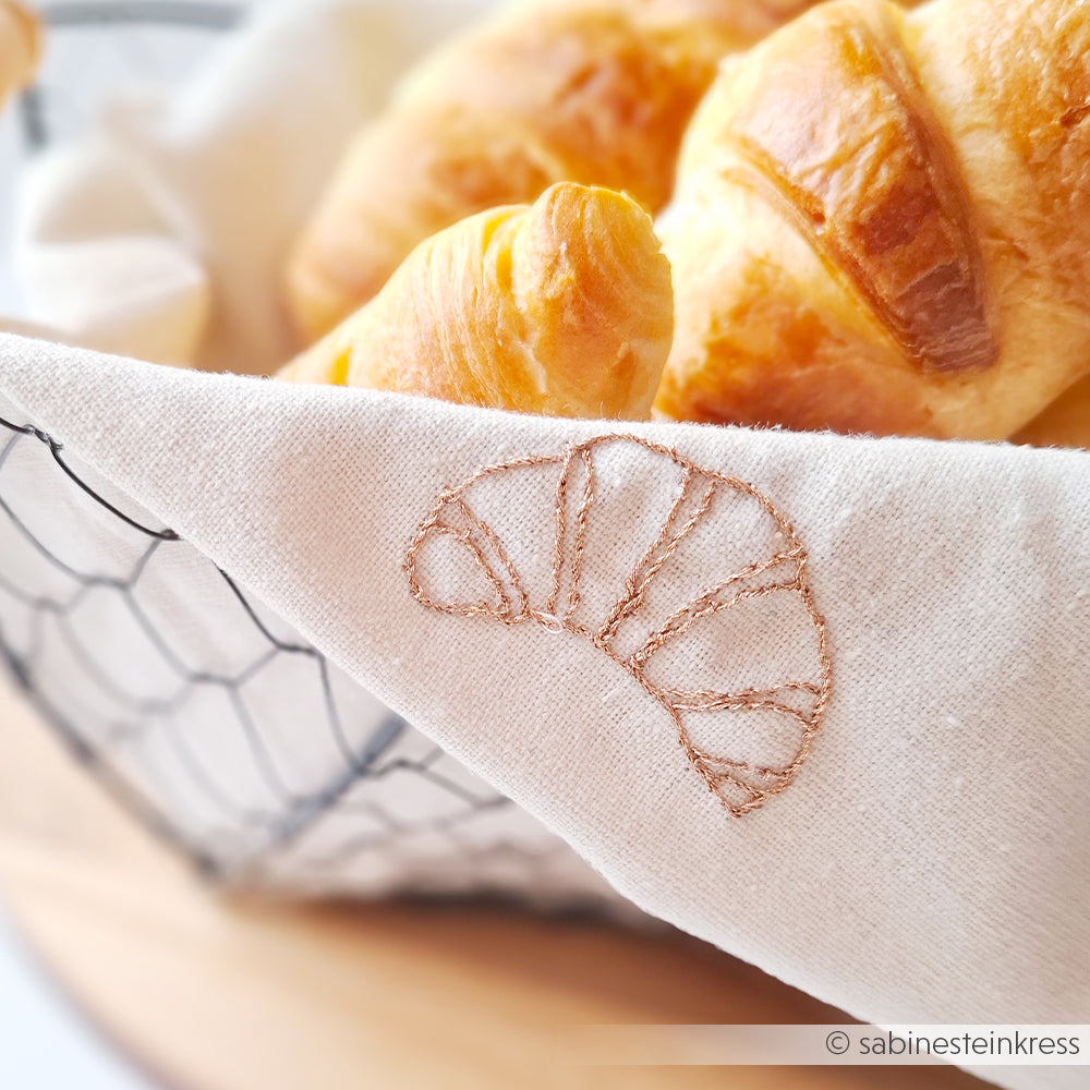 Ein Korb mit Croissants, ausgekleidet mit einem weißen Tuch mit der Croissant-Stickerei von Alexandra Renke, ist perfekt für Stickprojekte oder als Inspiration für Sticken-Vorlagen.