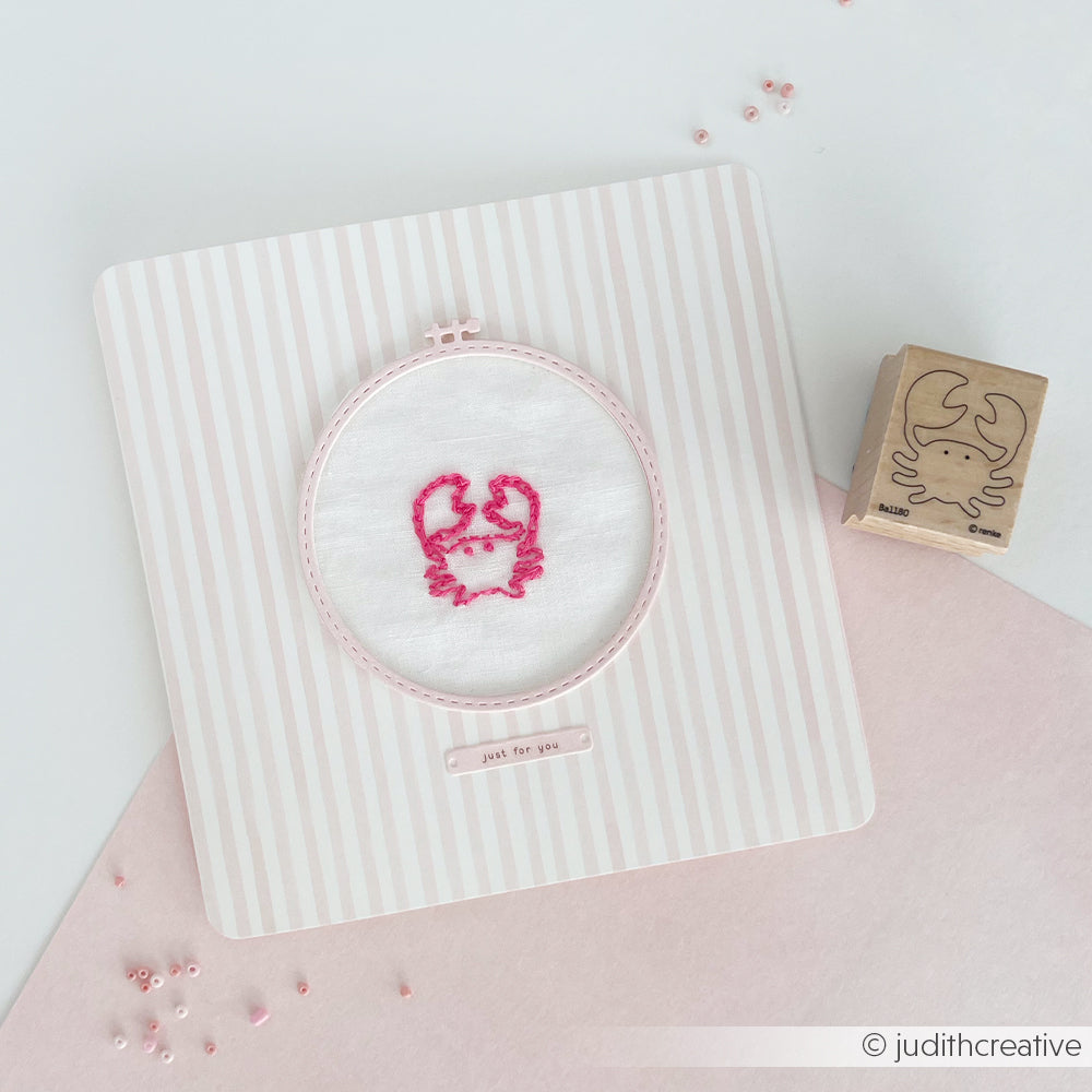 Der rosa Krabbenstickrahmen von Alexandra Renke auf einem gestreiften Hintergrund, mit einem Krabbenstempel (Stempelmotive für Stickerei) und rosa Perlen in der Nähe.