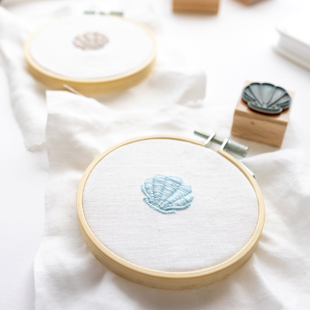 Zwei Stickrahmen von Alexandra Renke mit gestickten Muschelmotiven auf weißem Stoff, einer in Blau und einer in Beige - ideal für Stickprojekte oder als kreative Sticken-Vorlage - auf einer weißen Fläche ausgestellt.