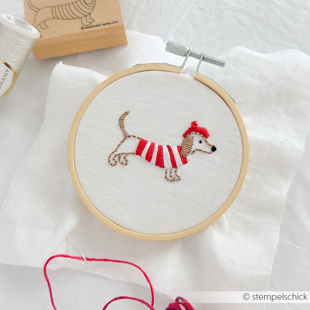 Die Stickerei von Alexandra Renke zeigt einen Dackel in einem rot gestreiften Pullover mit Hut, umrahmt auf weißem Stoff - perfekt für individuelle Geschenke oder als Inspiration für einzigartige Stickprojekte.
