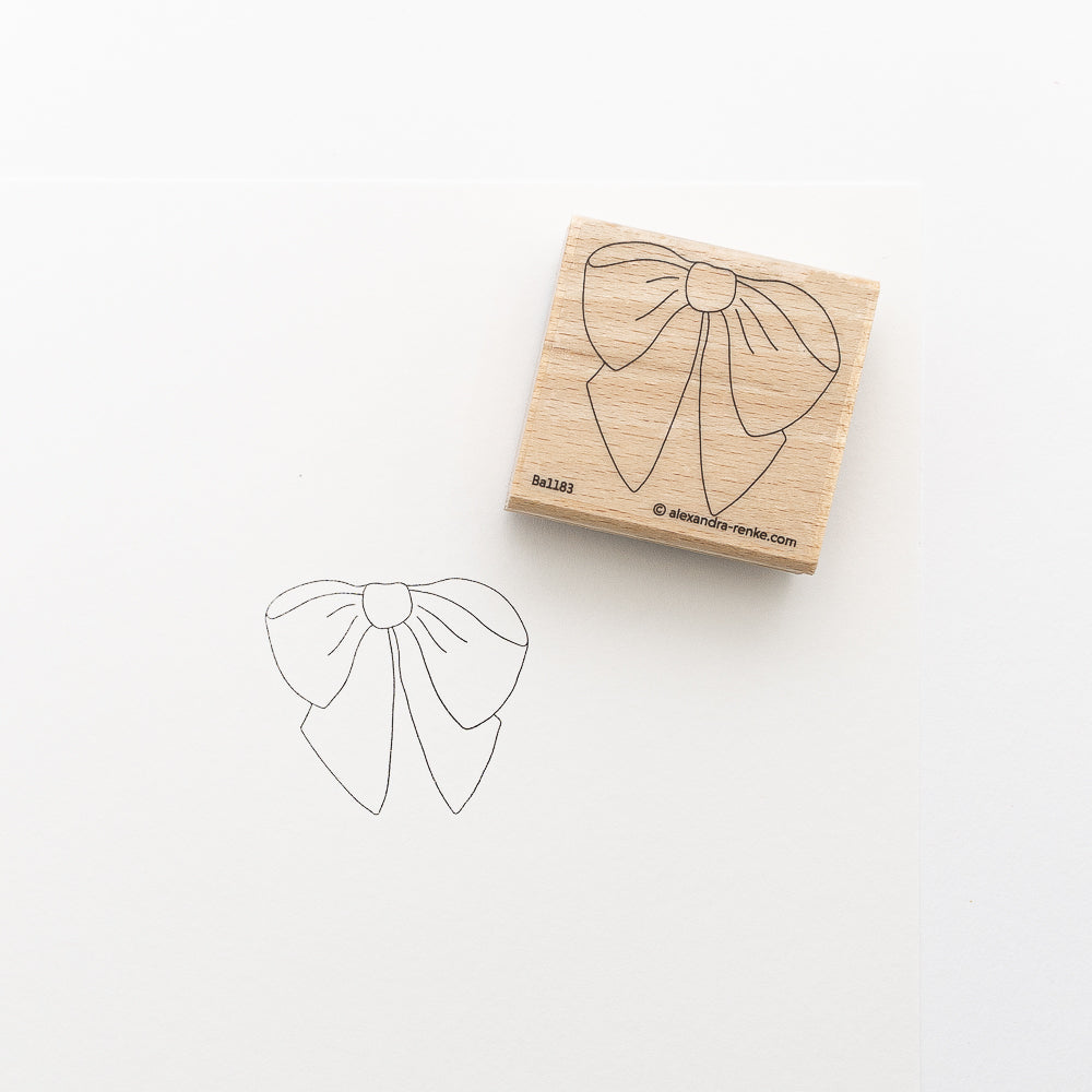Ein Holzstempel von Alexandra Renke mit Schleifen-Design liegt neben einem gestempelten Abdruck auf weißem Papier. Perfekt als Stickvorlage oder für Stempelfarbe auf Stoff geeignet.