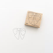 Ein Holzstempel von Alexandra Renke mit Schleifen-Design liegt neben einem gestempelten Abdruck auf weißem Papier. Perfekt als Stickvorlage oder für Stempelfarbe auf Stoff geeignet.