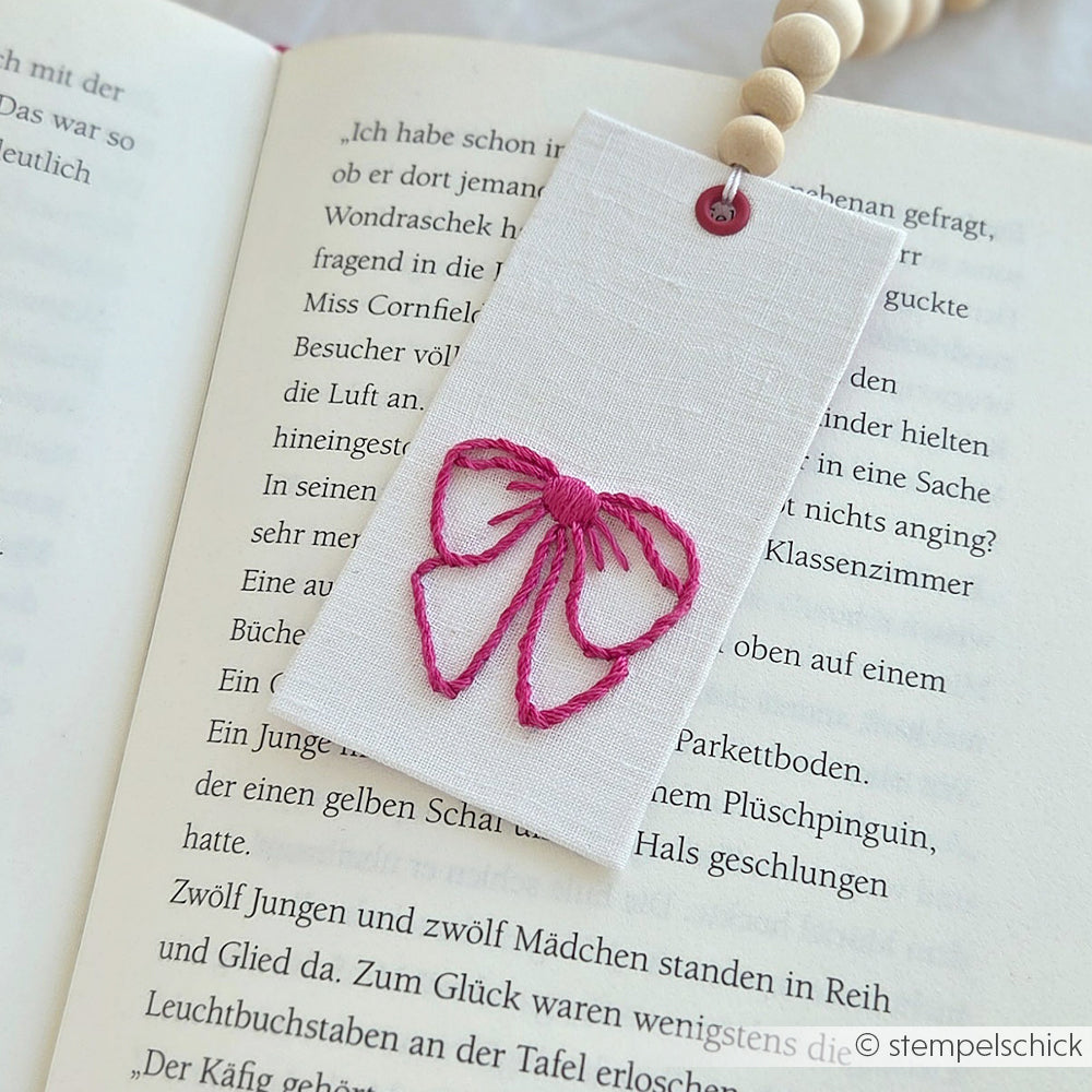 Auf einem aufgeschlagenen Buch mit deutschem Text ruht ein weißes Lesezeichen mit einer rosa gestickten Schleife, das mit einem Alexandra Renke Stickvorlage Stempel hergestellt wurde.
