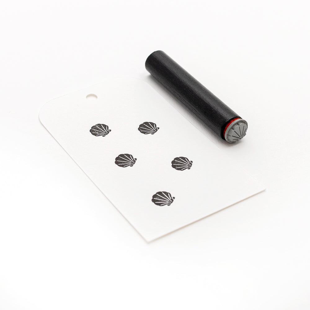 Ein schwarzer Alexandra Renke Mini Muschel Stempel und ein Anhänger mit sechs Muschelmotiven ruhen auf einem weißen Hintergrund, ideal für die Verwendung mit Ihrem Lieblings Stempelkissen oder Holzstempel.
