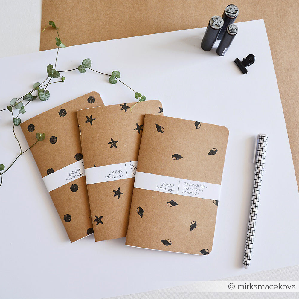 Drei braune Notizbücher von Alexandra Renke mit schwarzen Mustern, ein schwarzer Stift, Holzstempel und eine Weinrebe, angeordnet auf weißem Hintergrund.