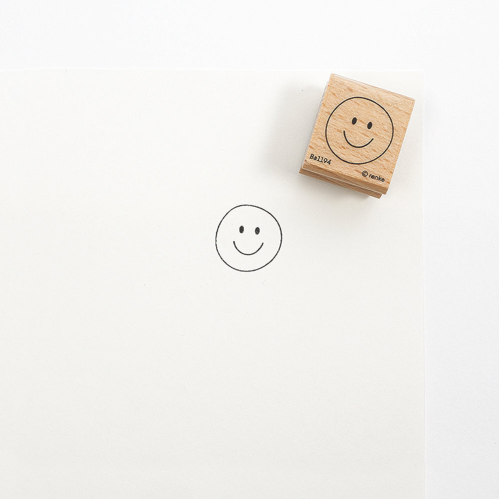 Ein Holzstempel mit einem Smiley liegt neben dem Abdruck auf einem leeren weißen Blatt Papier.