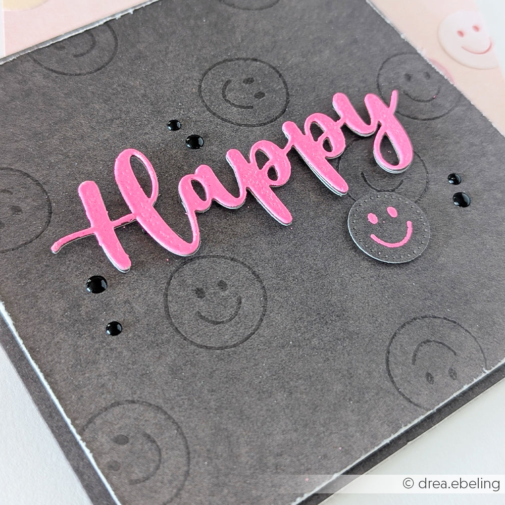 Handgefertigte Karte mit der Aufschrift „Happy“ in rosa Schreibschrift und fröhlichen Stempel-Smileys auf schwarzem Hintergrund mit kleinen schwarzen Punkten.
