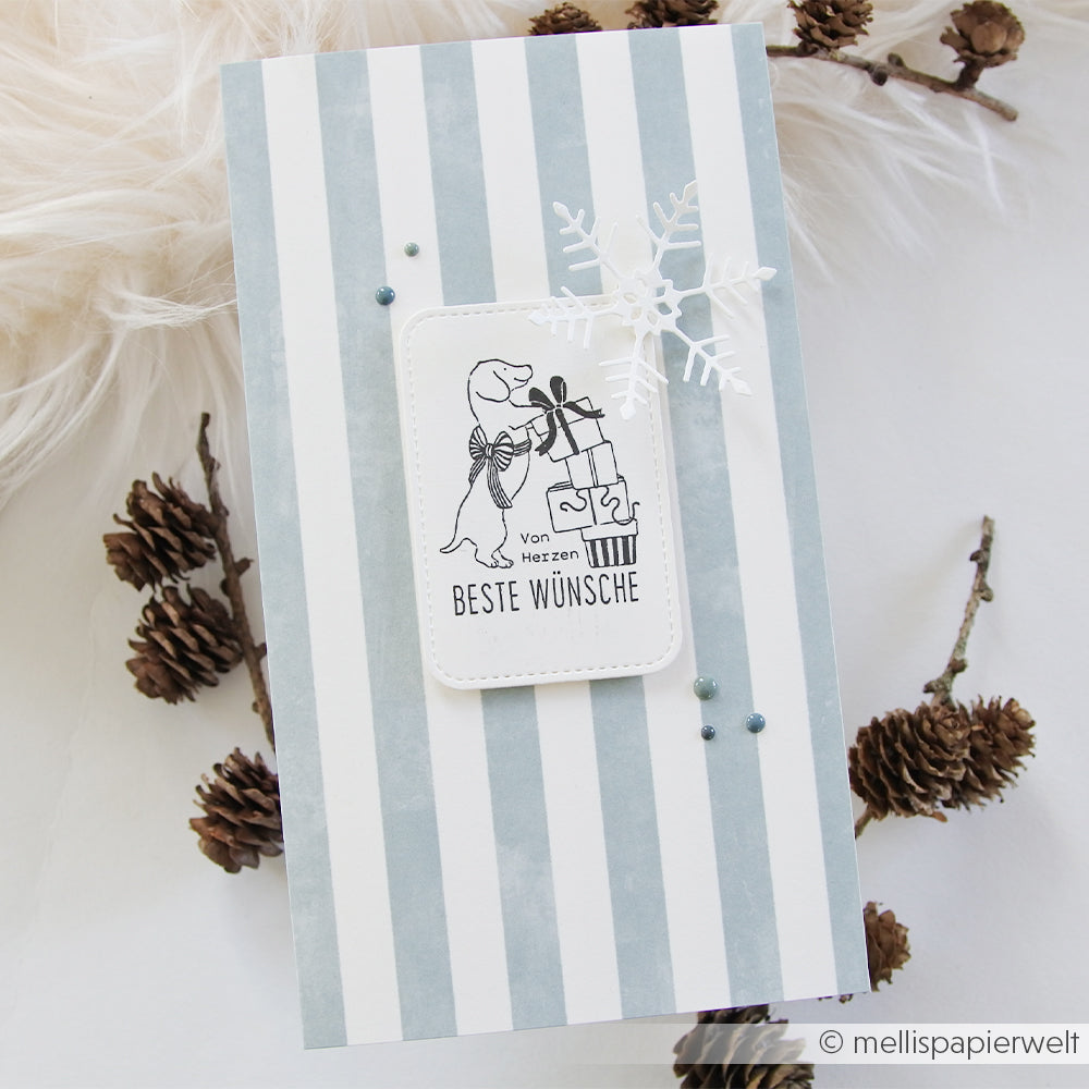 Handgefertigte Karte mit gestreiftem Hintergrund, einem Dackel, der Geschenke hält, und einem Holzstempel mit Schneeflocke.