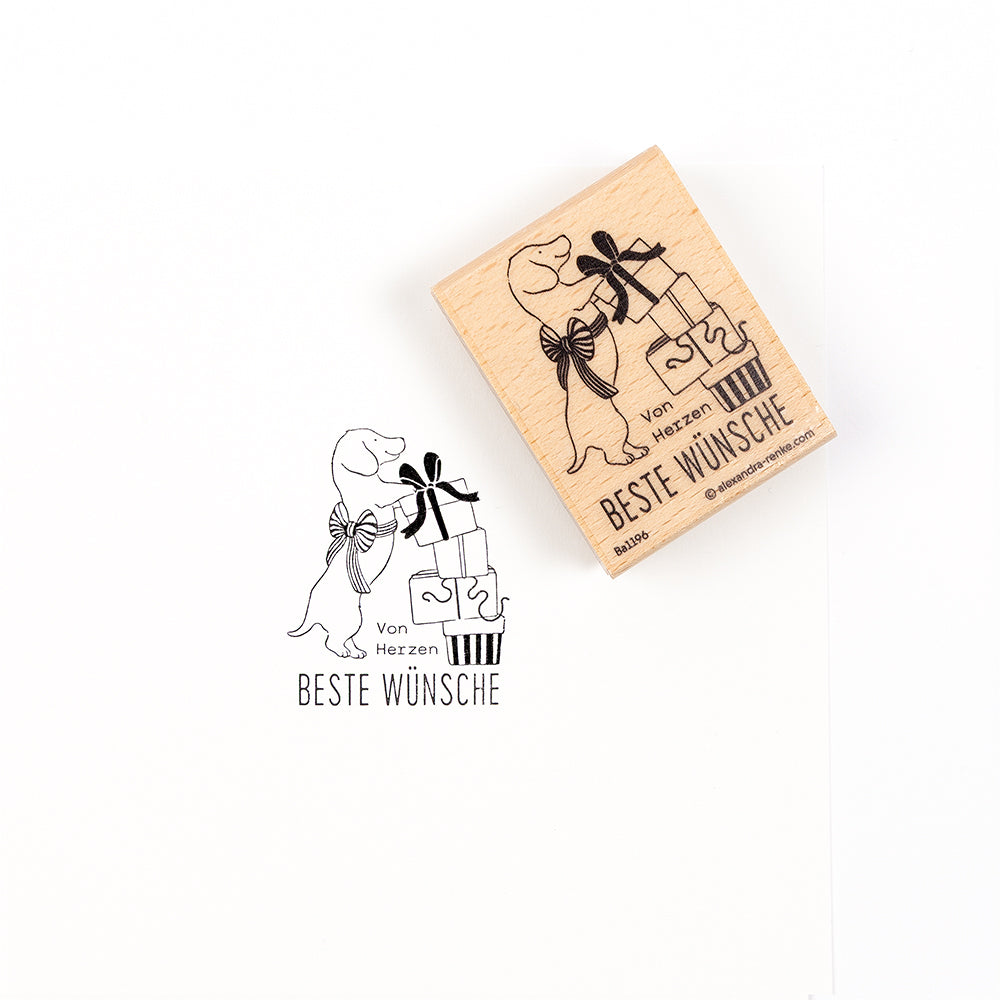 Ein Holzstempel und sein Abdruck, der einen Dackel mit Geschenkpaketen und den deutschen Text „Von Herzen BESTE WÜNSCHE“ zeigt.