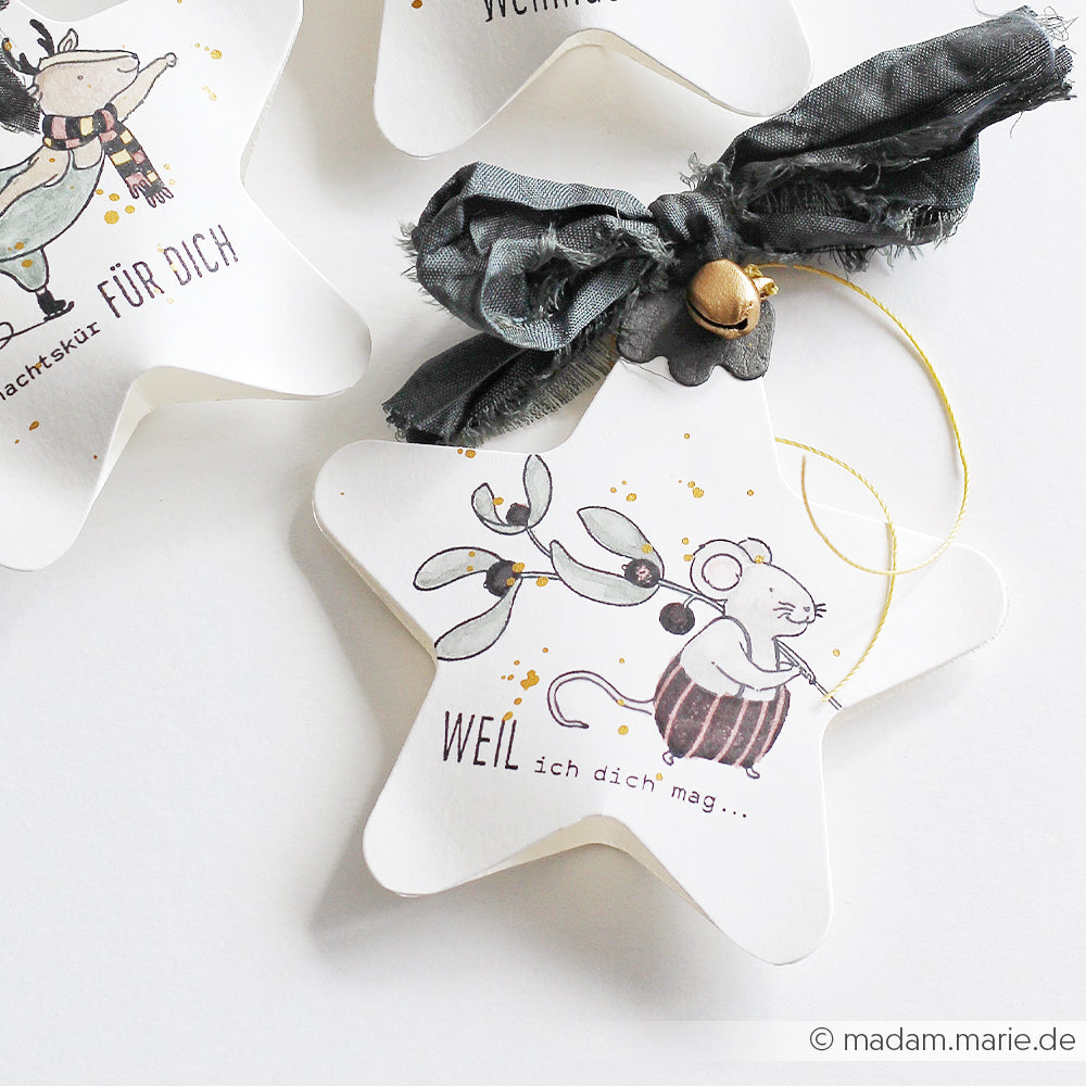 Sternförmige Keramikornamente mit einer Mausillustration, die mit einem Holzstempel angefertigt wurde, mit einem deutschen Text beschriftet und mit einem grauen Band und einem Glöckchen verziert.