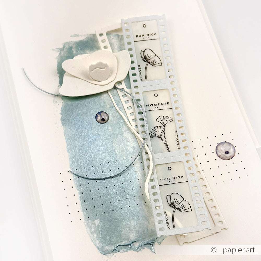 Eine handgefertigte Karte von Alexandra Renke mit weißer Mohnblume, Filmstreifen, Blattskizzen, blauen Metallic-Farbakzenten und einzigartigen Stempelgummi-Details.