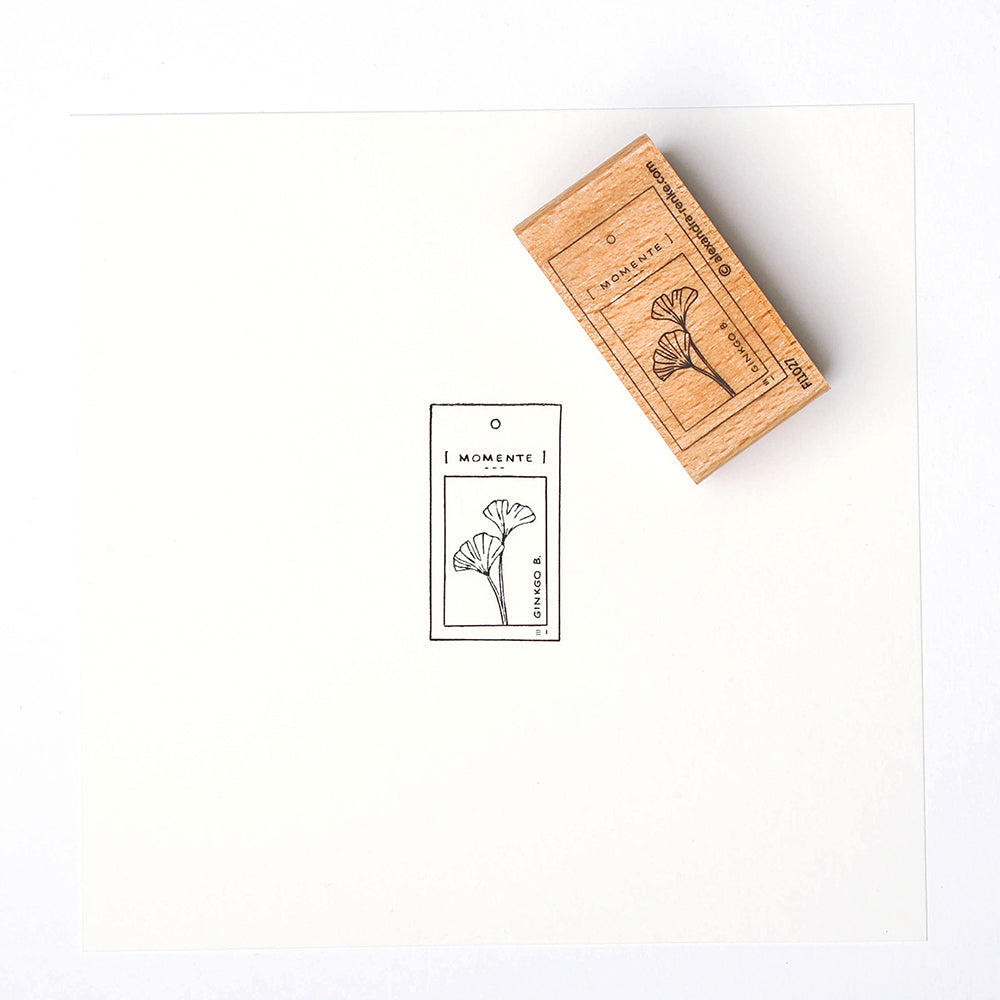 Ein Alexandra Renke Holzstempel und sein gestempeltes Bild einer Blume mit dem Text "MOMENTE" auf weißem Papier.