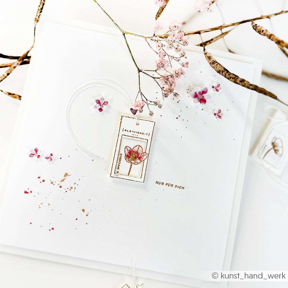 Eine zarte, handgefertigte Karte von Alexandra Renke mit rosafarbenen Blumen, goldenen Akzenten, einem Blumenanhänger auf weißem Grund und einem eleganten Krokus-Stempel-Motiv für eine charmante Note.