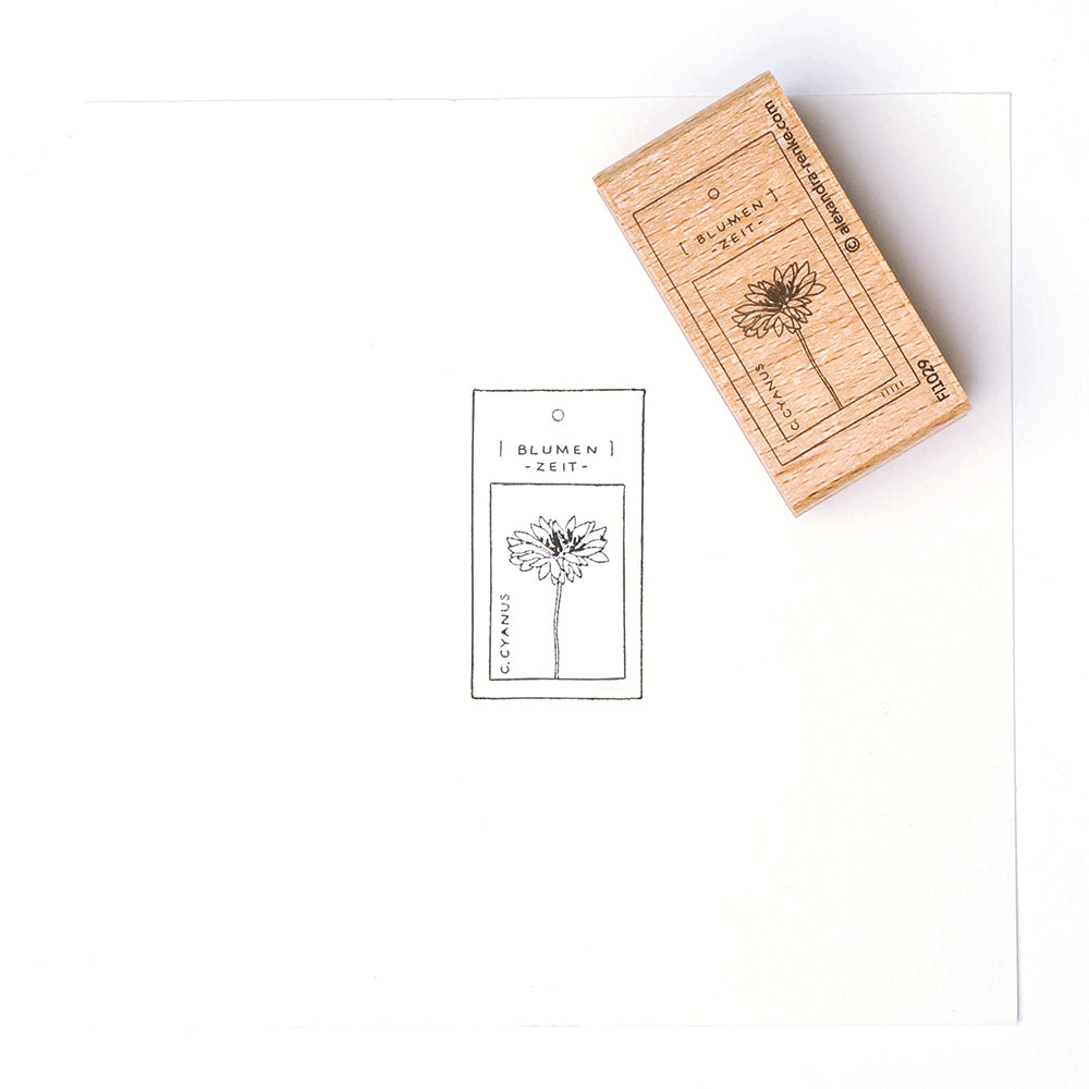 Ein Holzstempel von Alexandra Renke erzeugt einen Blumenabdruck mit schwarzer Tinte auf einem BLUMEN ZEIT-Anhänger, der mit einem Stempelkissen hergestellt wurde.