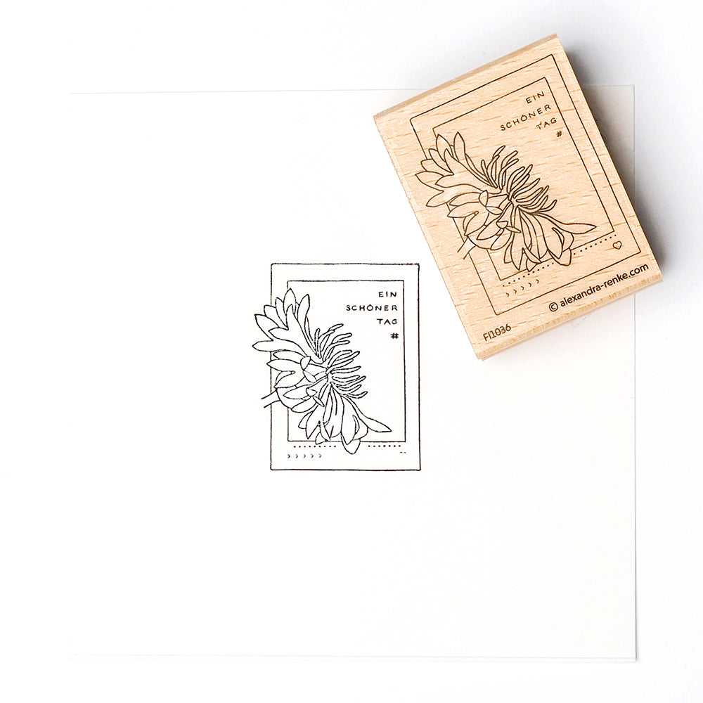 Ein Holzstempel von Alexandra Renke und sein Abdruck mit einer Blume und dem Text "Ein schöner Tag" auf weißem Papier - ideal für kreative Papierprojekte.