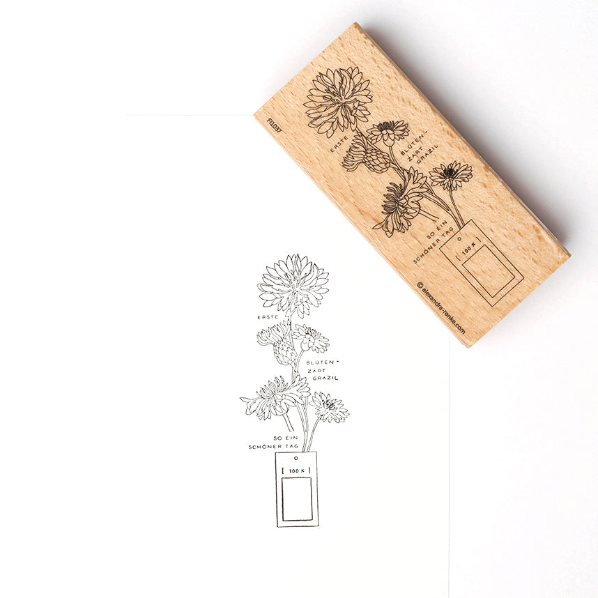Ein Alexandra Renke Holzstempel und sein gestempeltes Bild von Blumen in einer rechteckigen Vase, mit dem deutschen Text "Kornblumen grazil", gezeigt auf weißem Papier.